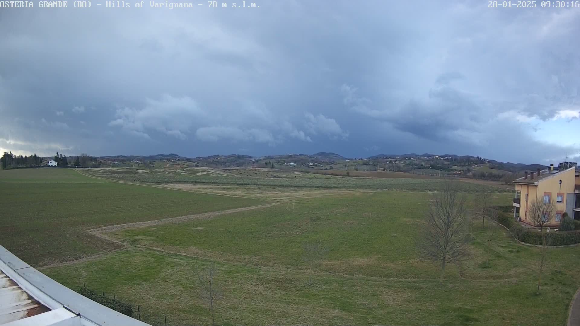 Under a cloudy sky, a wide expanse of flat, green farmland stretches to rolling hills in the distance, with a few buildings visible near the bottom right corner.