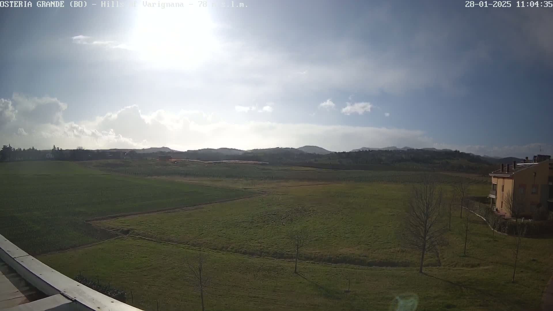 A partly cloudy day reveals a sprawling green field, dotted with leafless trees, leading to rolling hills in the distance, with a lone building visible on the lower right.