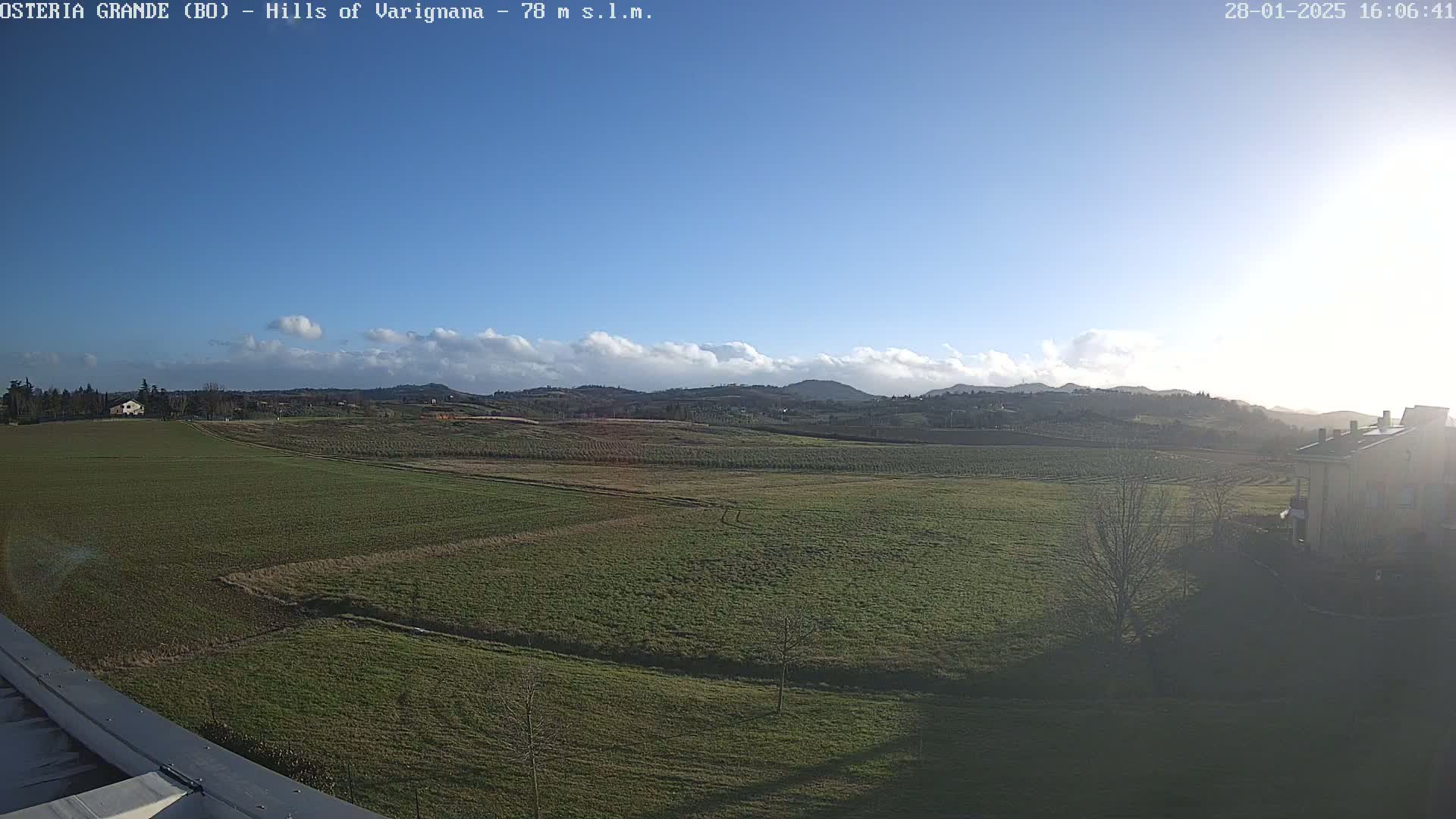 A partly cloudy, sunny day reveals a sprawling green field, dotted with trees and buildings, extending to distant rolling hills.