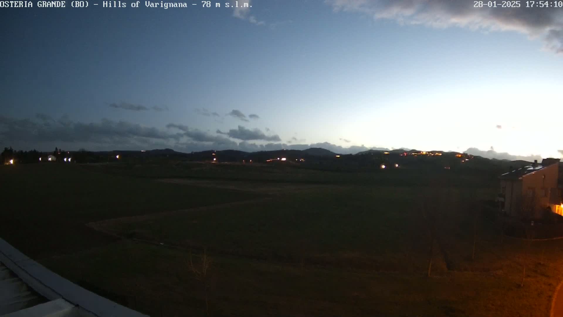 A twilight view of a dark green field, with distant hills and a few lights visible under a partly cloudy sky.