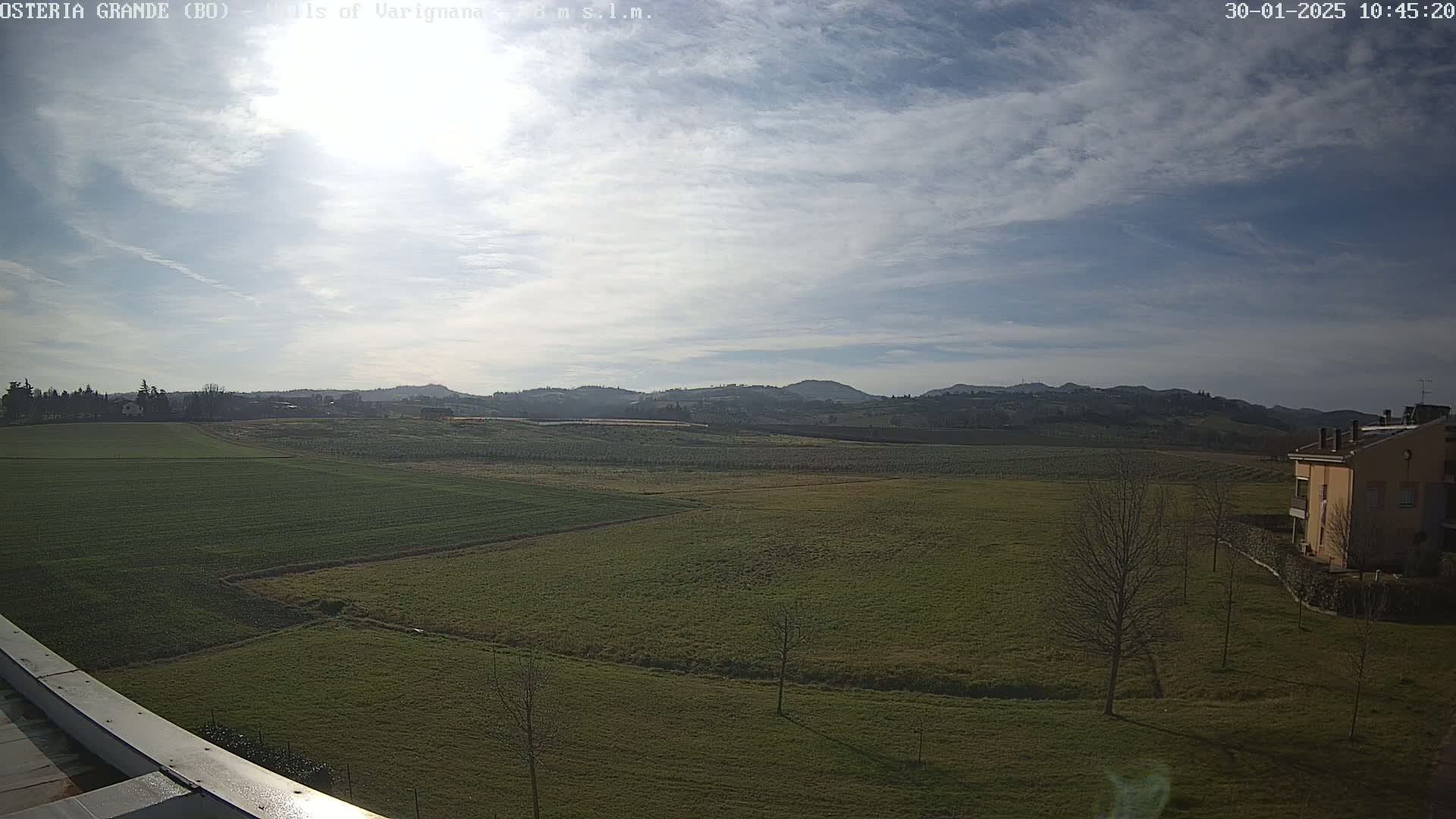 A sunny day reveals a sprawling green field, bordered by a few buildings and trees, with rolling hills in the distance under a partly cloudy sky.