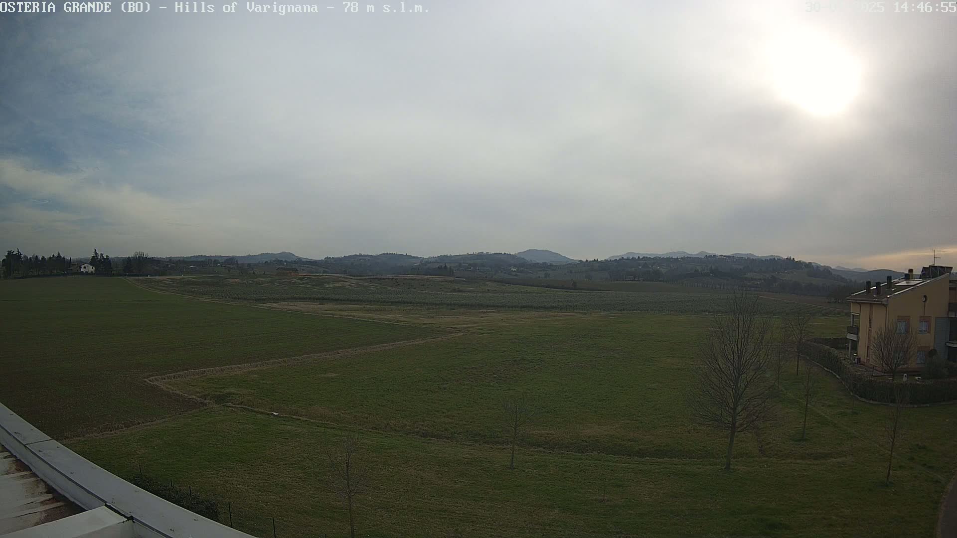 A mostly sunny day reveals a wide expanse of green fields, a few sparsely-leafed trees, distant hills, and a tan building in the bottom right corner.