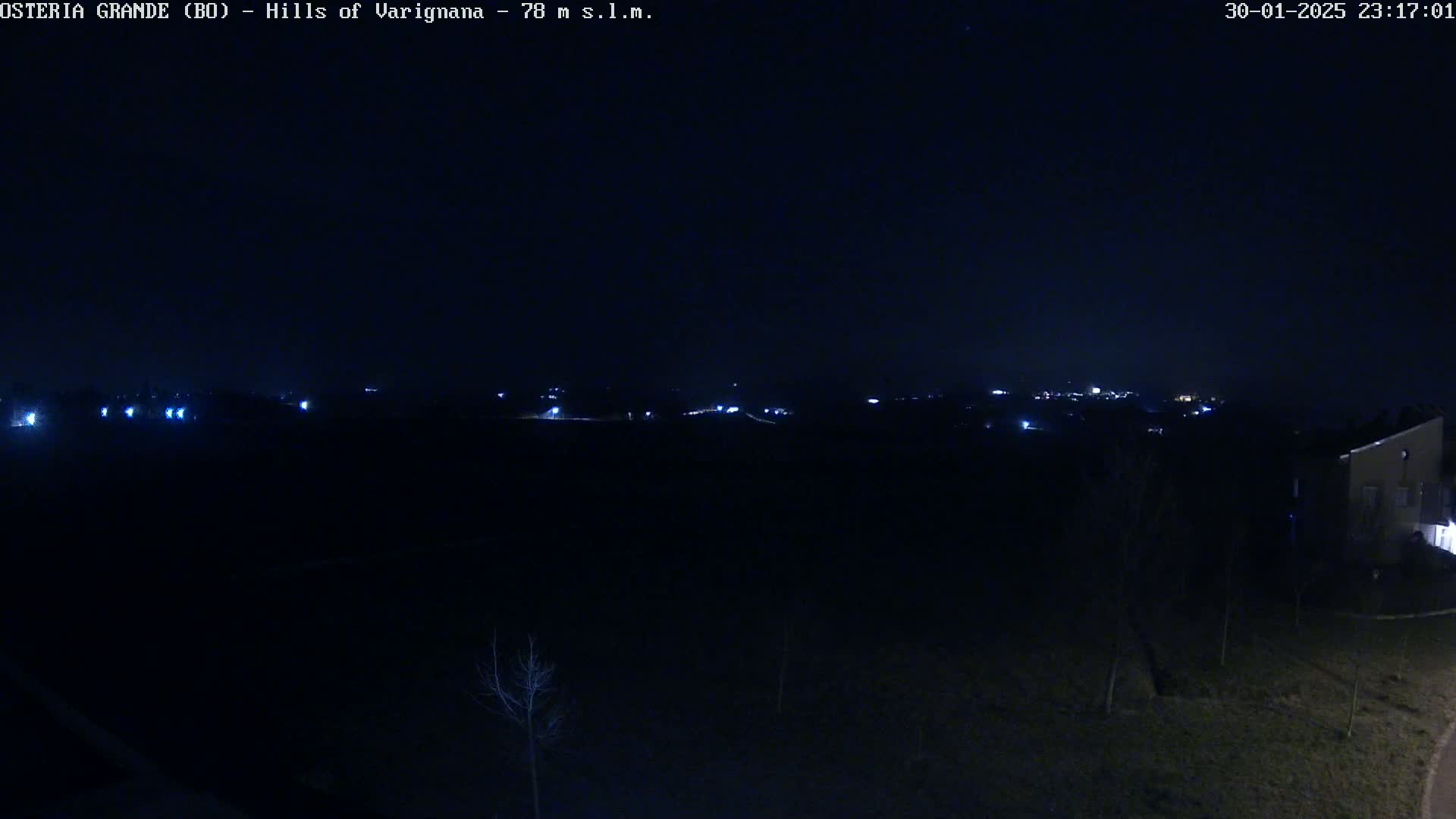 A nighttime, dark view of a sparsely lit landscape with a few distant lights and a building partially visible in the lower right corner.