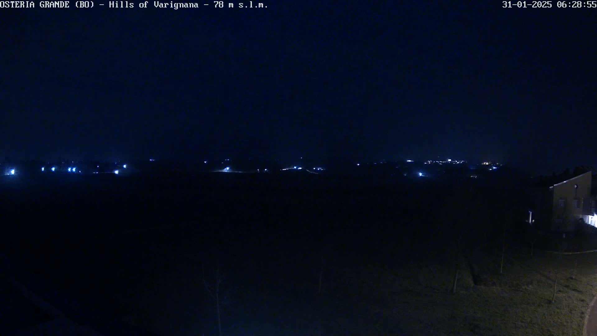 A dark, nighttime landscape shows a distant line of lights on a hill, with a few closer lights visible in the foreground.