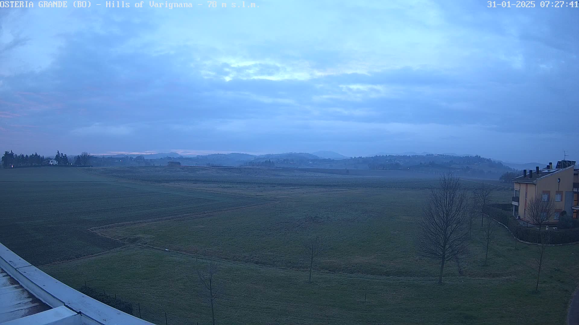A wide, flat expanse of farmland under a pale blue, cloudy sky stretches towards distant, hazy hills.