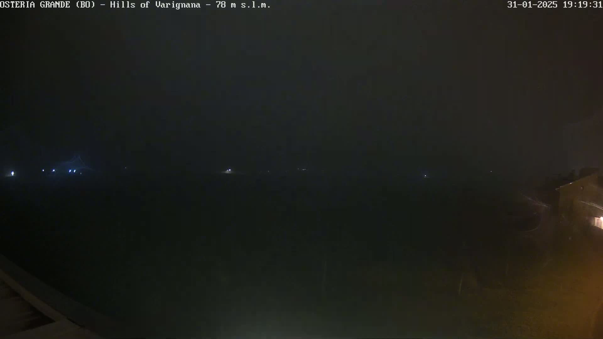 A dark, nighttime landscape is shrouded in fog or mist, with a few faint lights visible in the distance.