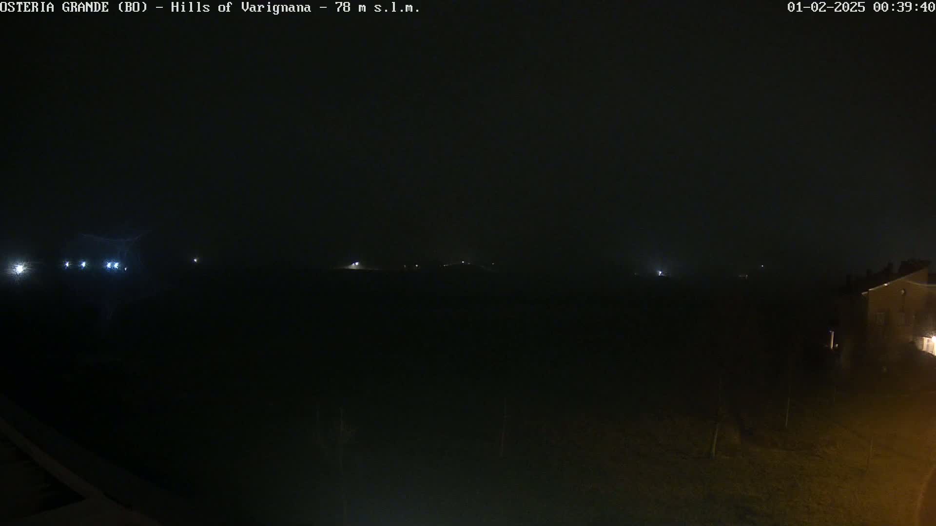 A dark, nighttime landscape is visible, with a few faint lights in the distance and a building on the lower right.