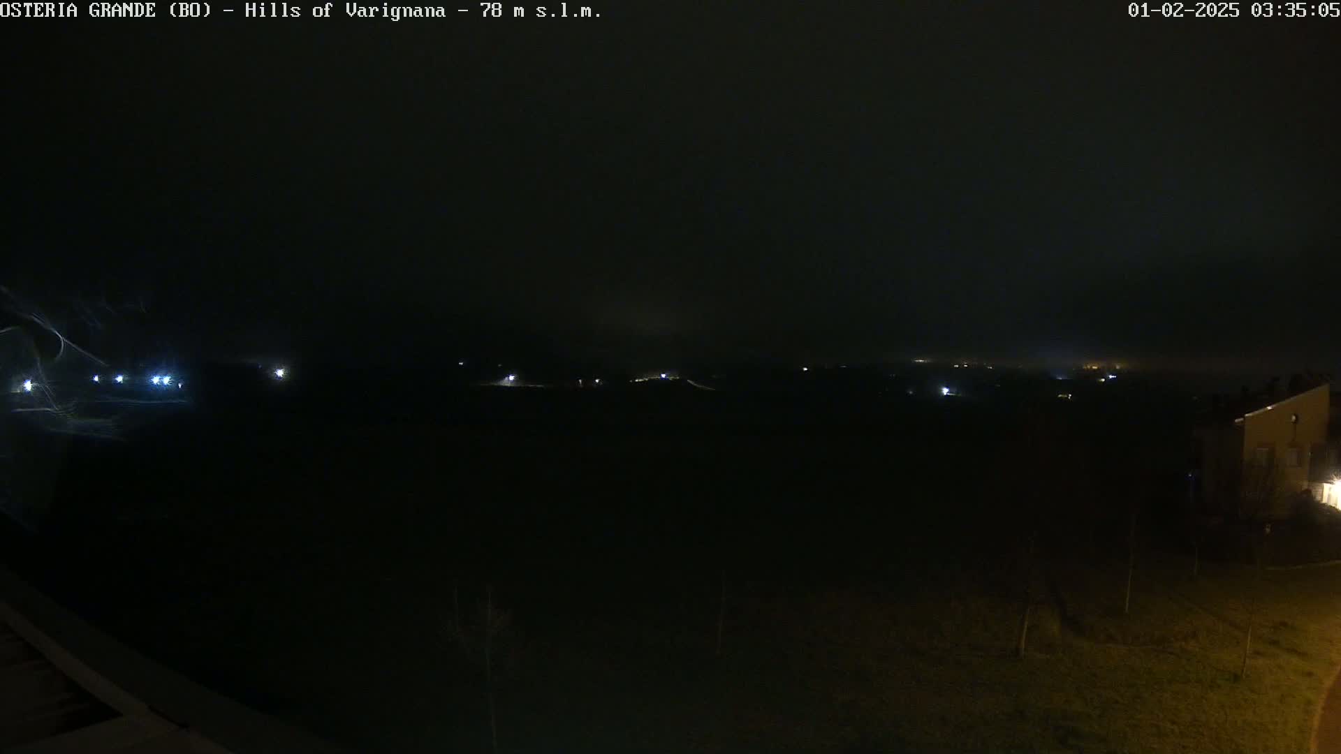 A nighttime, foggy view shows a dark landscape with faint lights in the distance and a building on the right.
