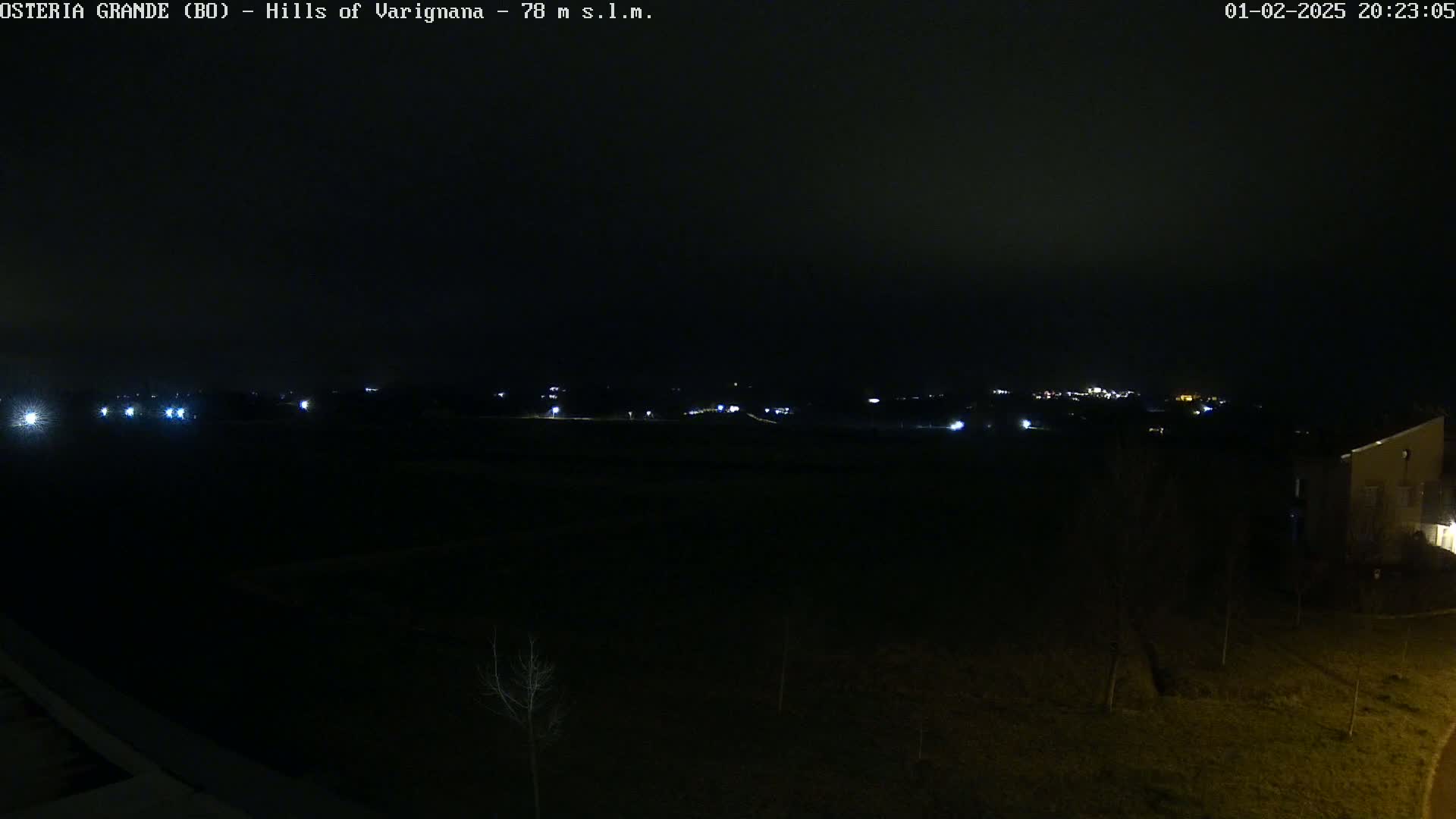 A nighttime, dark view of a mostly flat landscape with a few distant lights and a building visible in the bottom right corner under a cloudy sky.