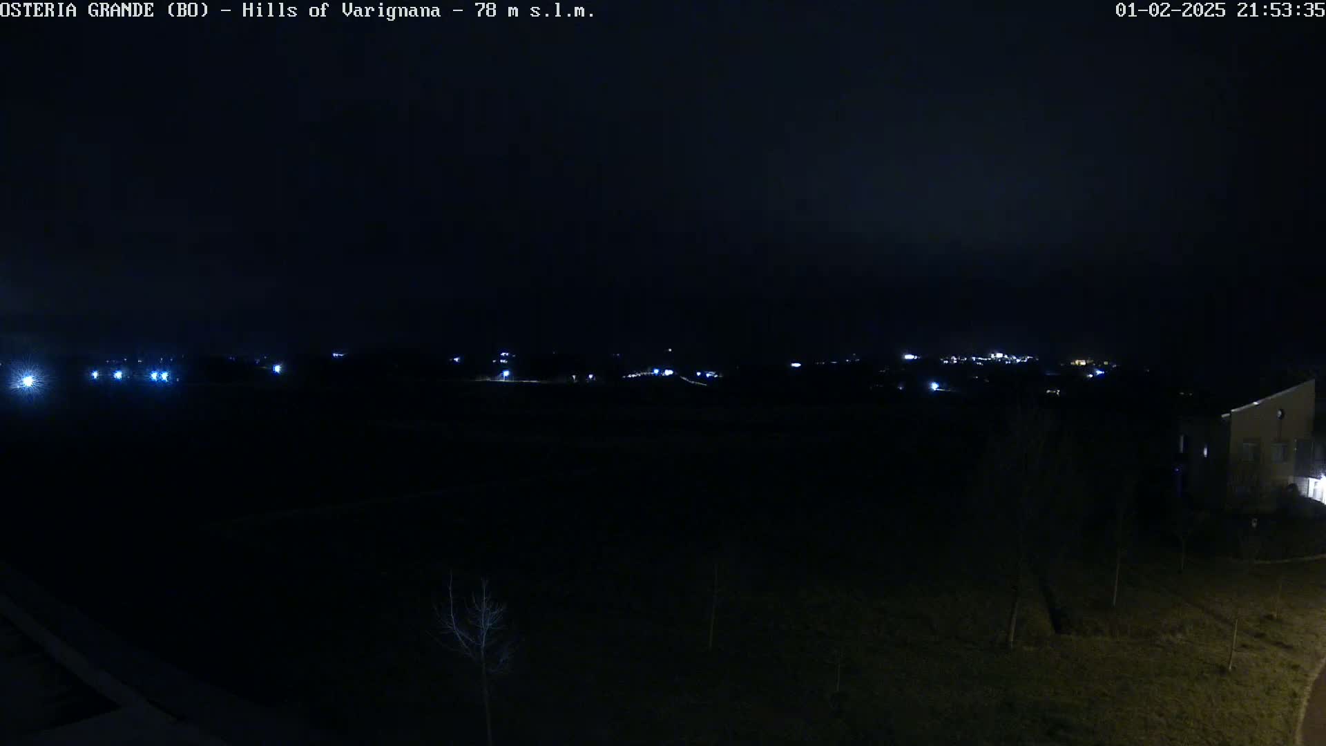 A nighttime, dark view of a sparsely lit landscape with a few distant lights and a building in the bottom right corner.