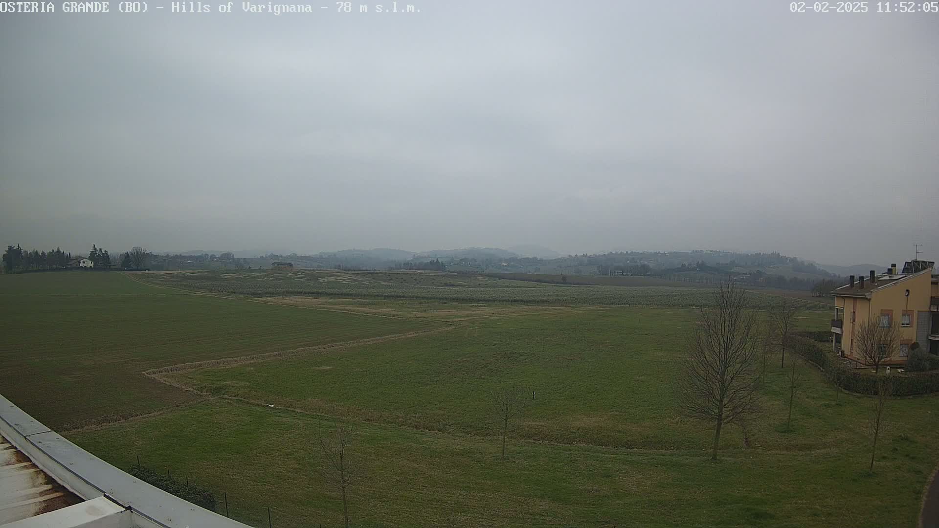 A wide, overcast view of a flat, green field with a few bare trees and a building in the lower right, and rolling hills in the distance.