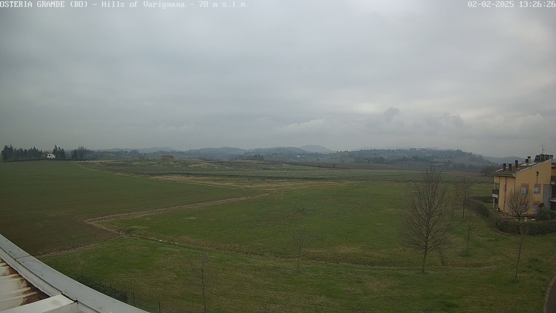 A wide, overcast view of a flat, green field with a few trees and buildings in the distance, under a gray sky.