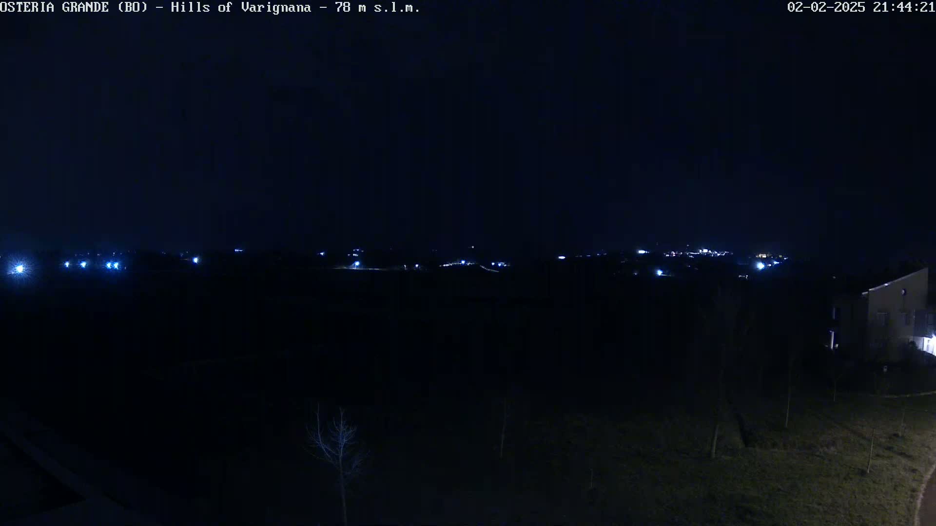 A dark, nighttime landscape shows a distant line of faint lights, likely from a town or village, with a few lights closer to the camera and a building at the lower right.