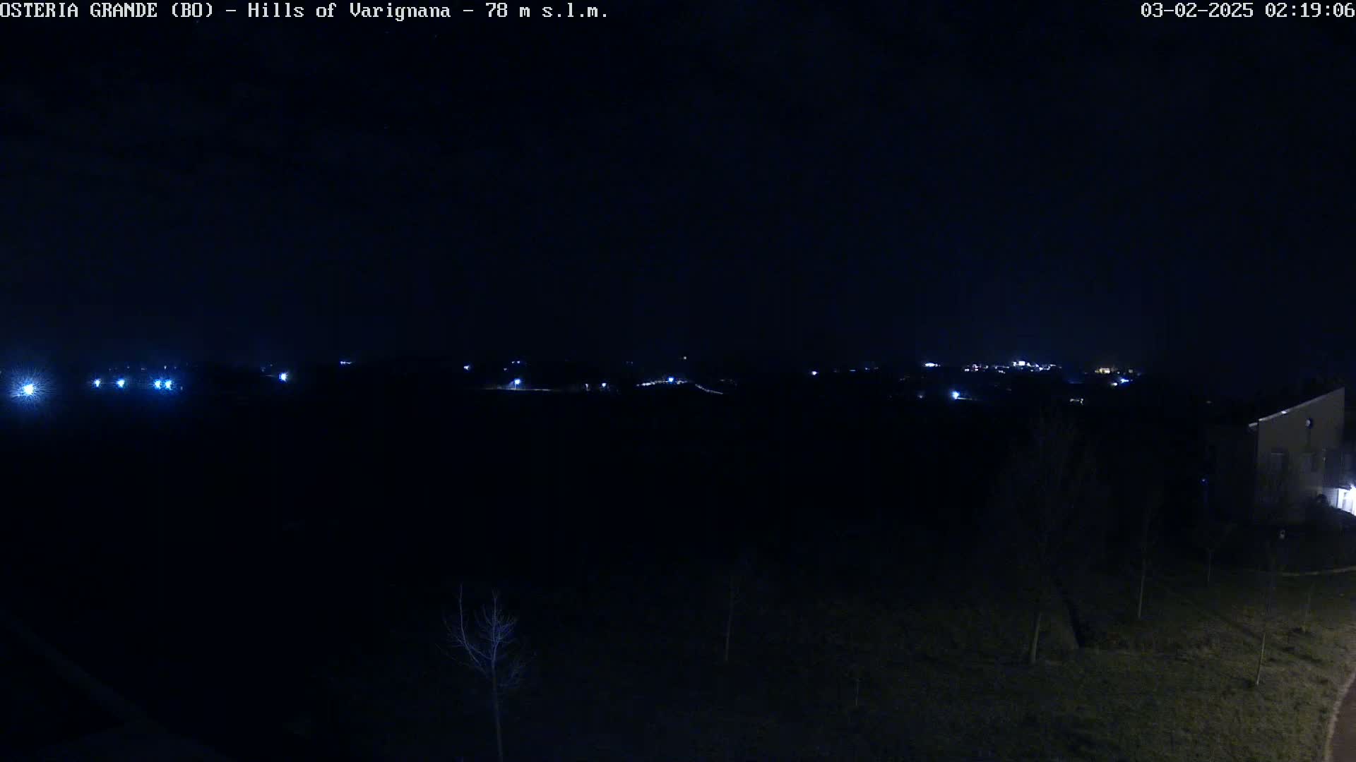 A nighttime, dark landscape shows a distant line of lights on a hill, with a few lights closer to the foreground and a building visible on the lower right.