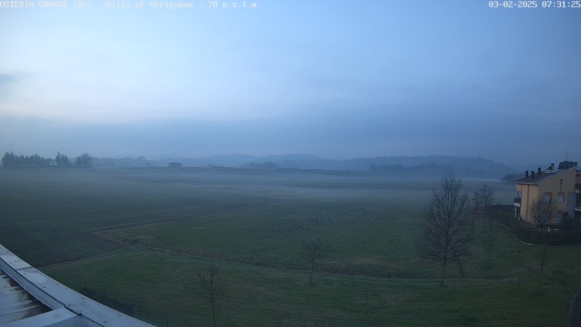 A foggy, early morning view of a flat, green field extending to distant, low hills under a pale blue sky.