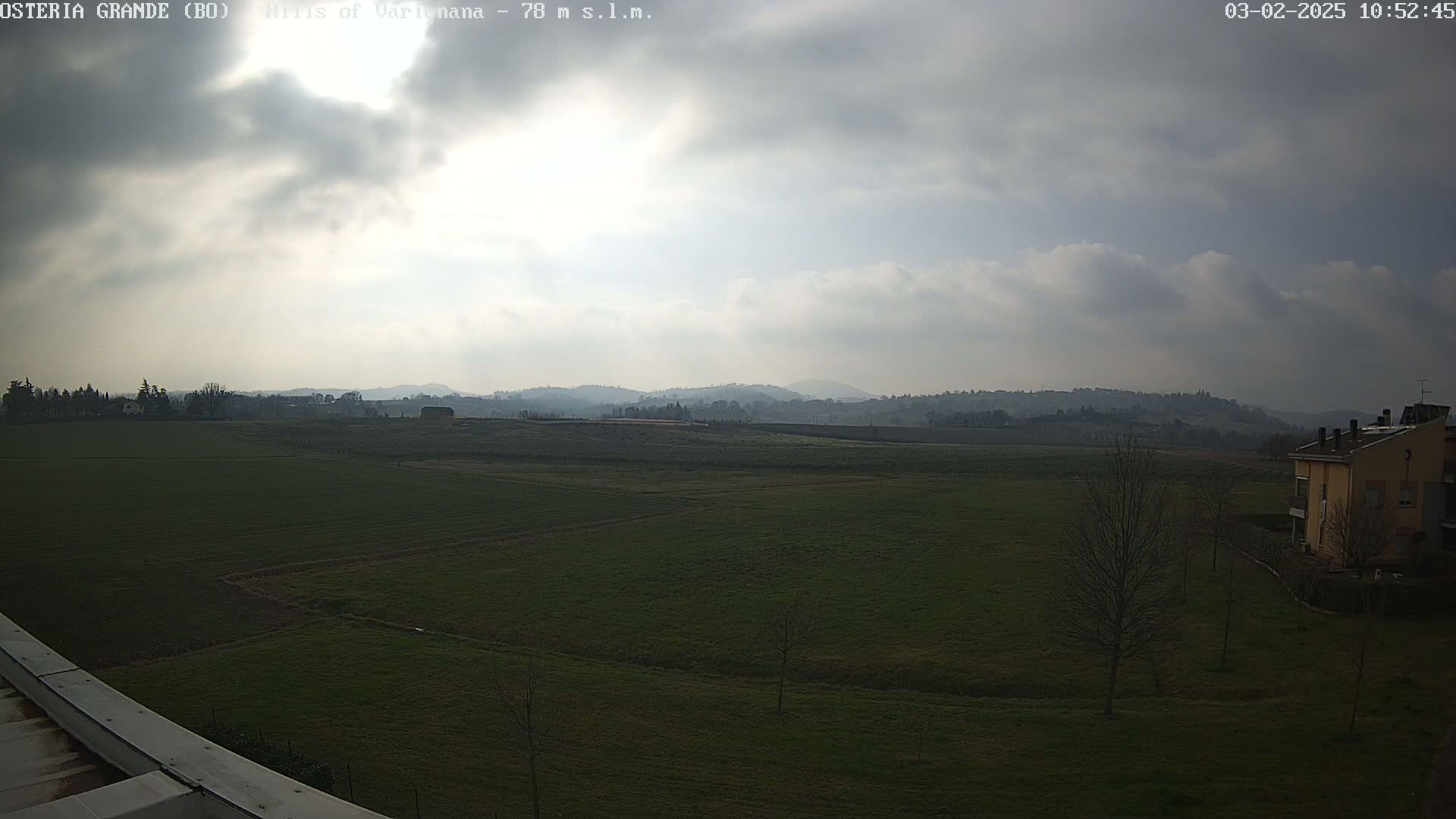 A mostly cloudy day reveals a flat, green field extending to rolling hills in the distance, with a few buildings visible near the bottom right.