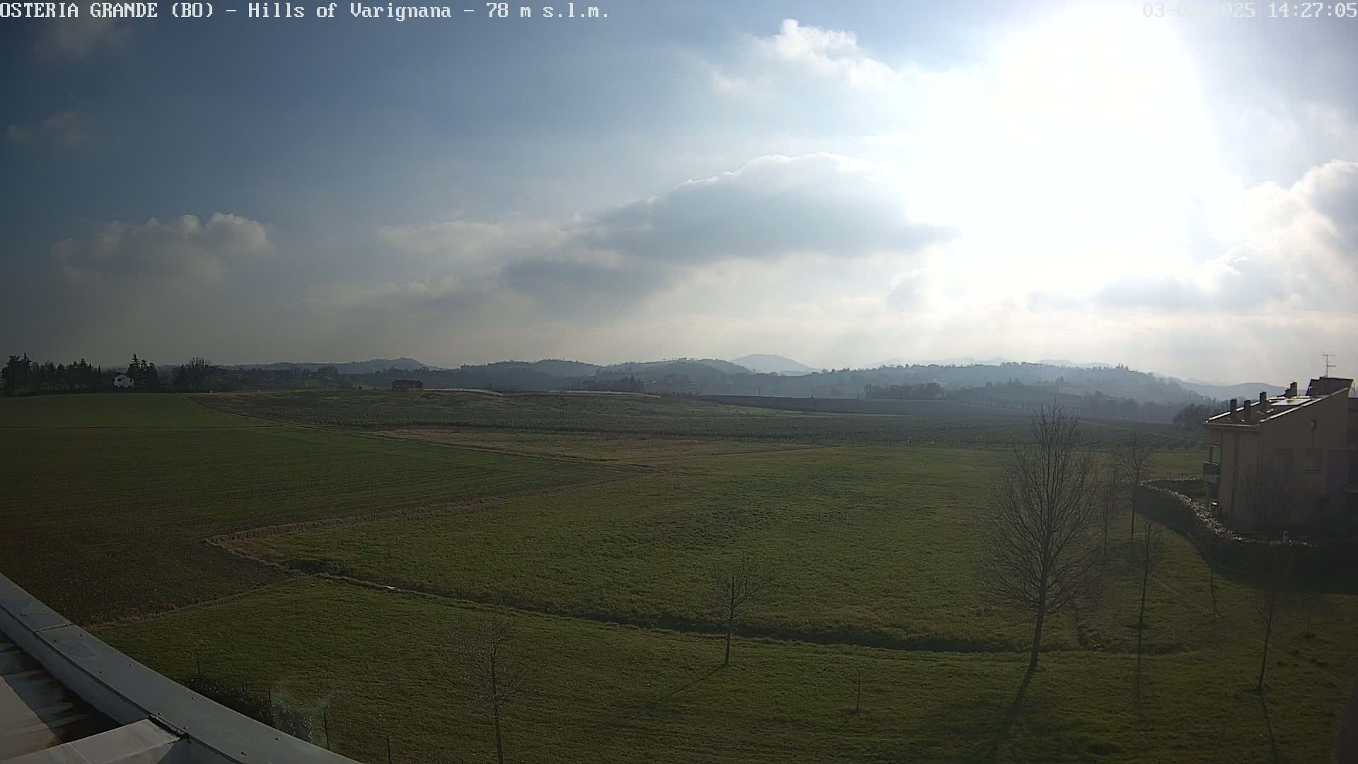 A partly cloudy, sunny day reveals a vast expanse of verdant farmland, punctuated by a few sparse trees and a building in the lower right corner, all nestled beneath rolling hills in the distance.