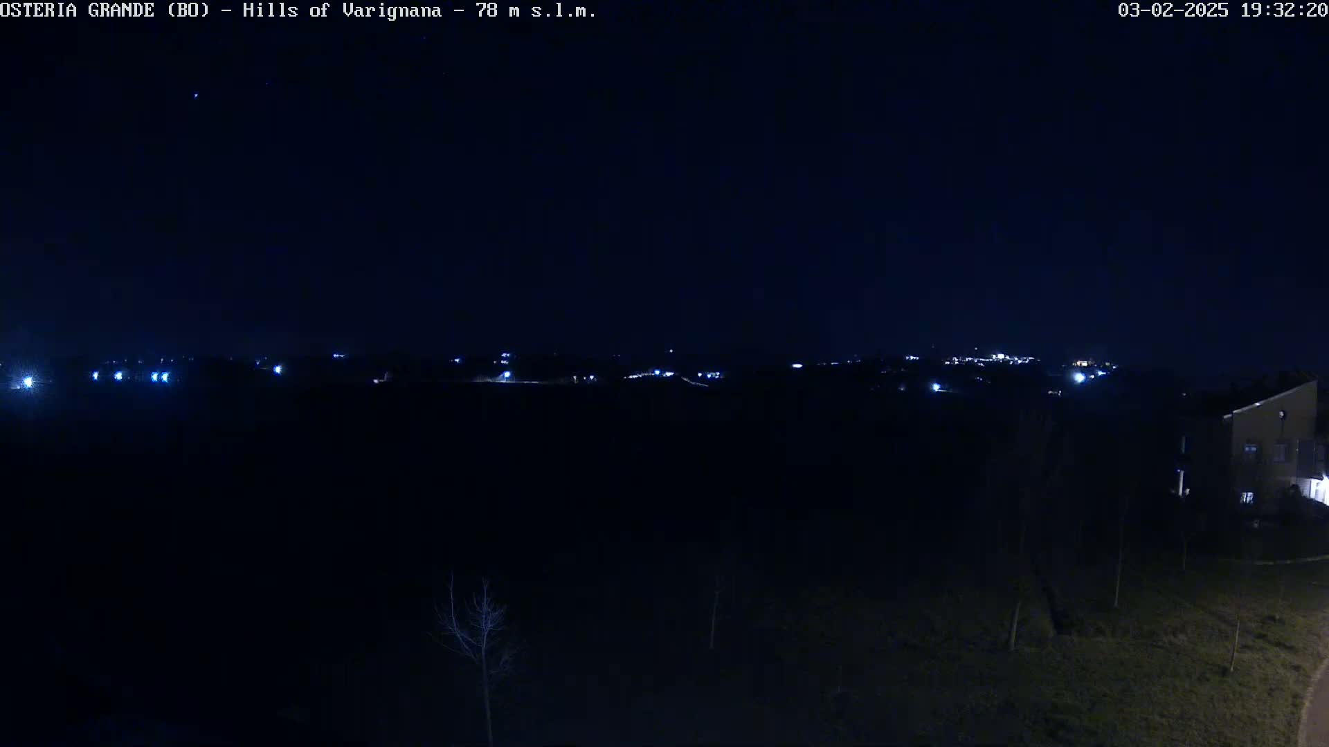 A nighttime, long shot of dimly lit hills and a small cluster of buildings in the distance under a clear, dark sky.