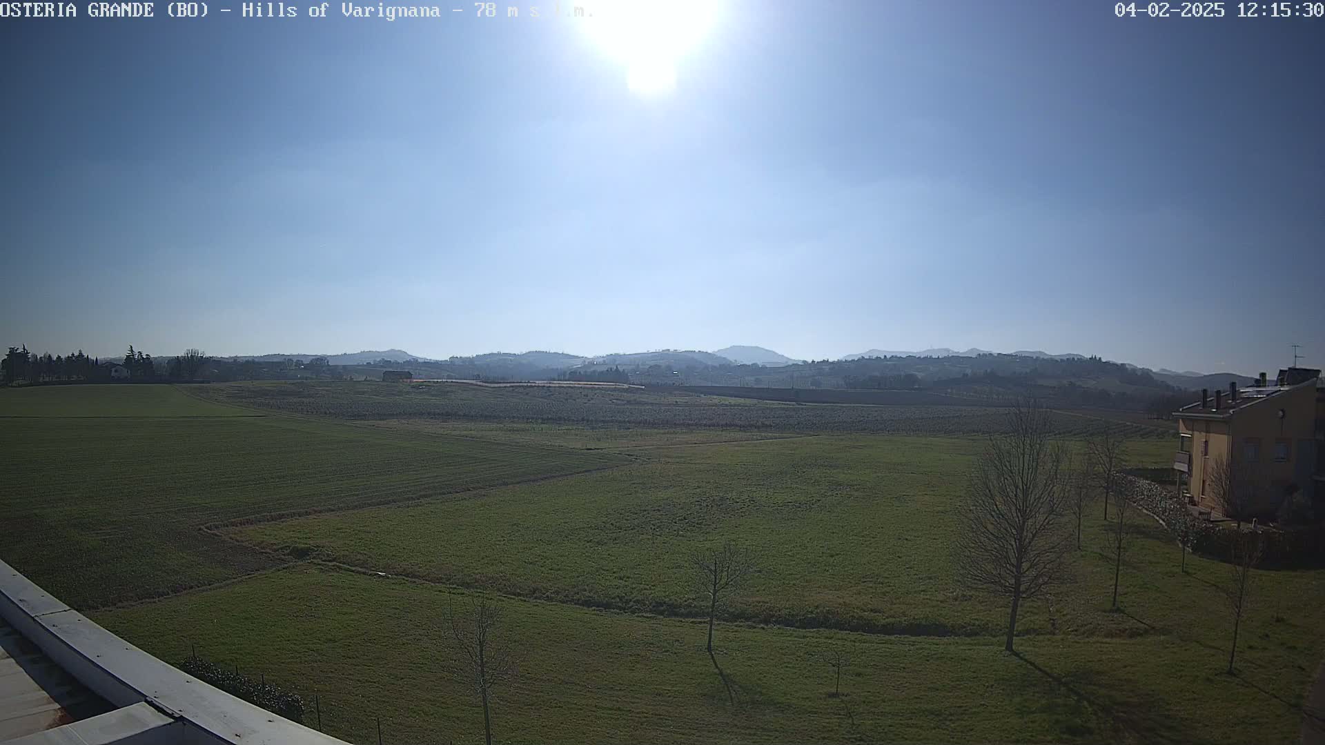 A sunny day reveals a vast expanse of green fields, dotted with bare winter trees, stretching towards a range of distant hills under a clear blue sky.