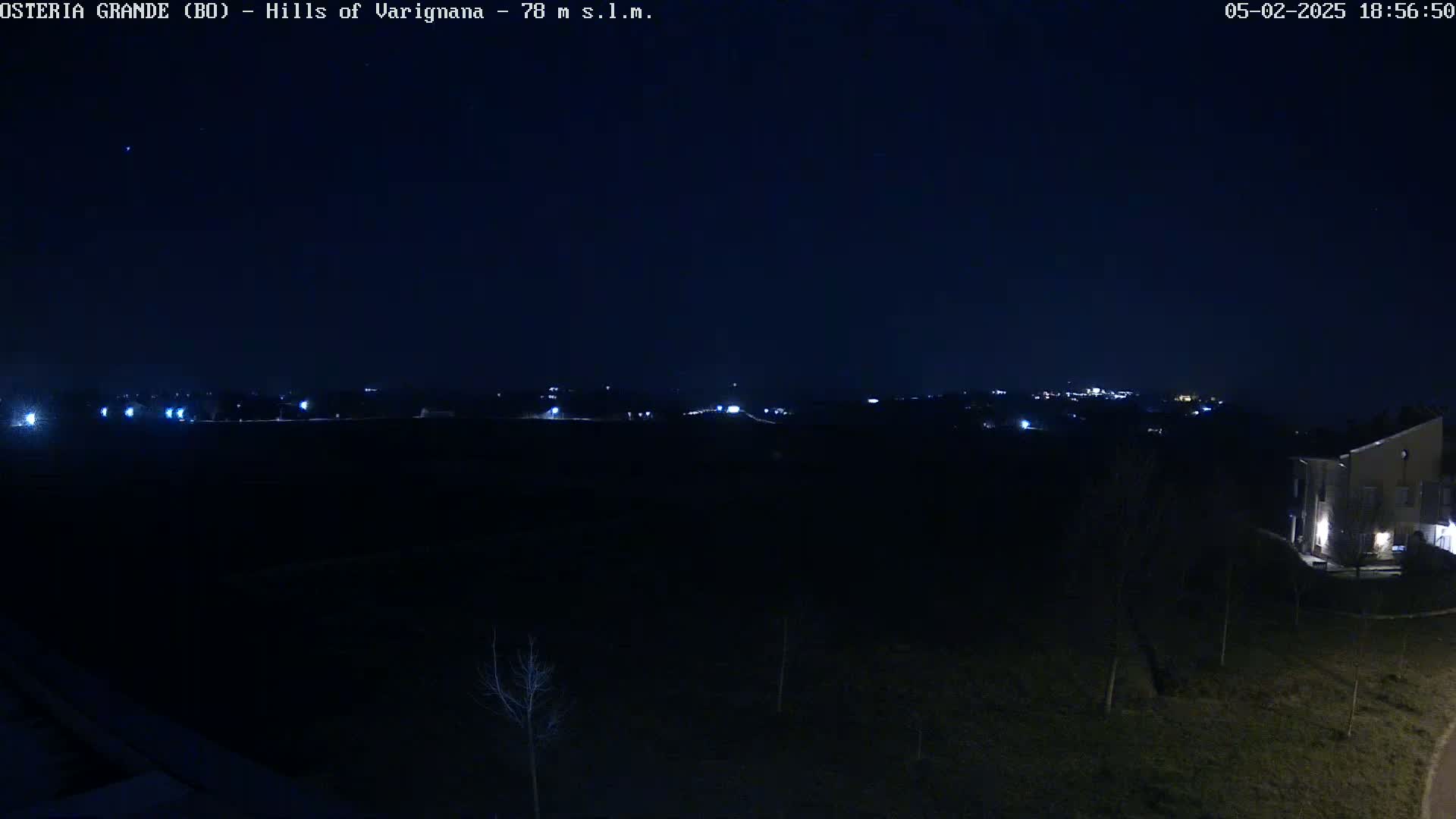 A nighttime long shot shows a dark landscape with a few distant lights, a building in the lower right corner, and a mostly clear, dark sky.