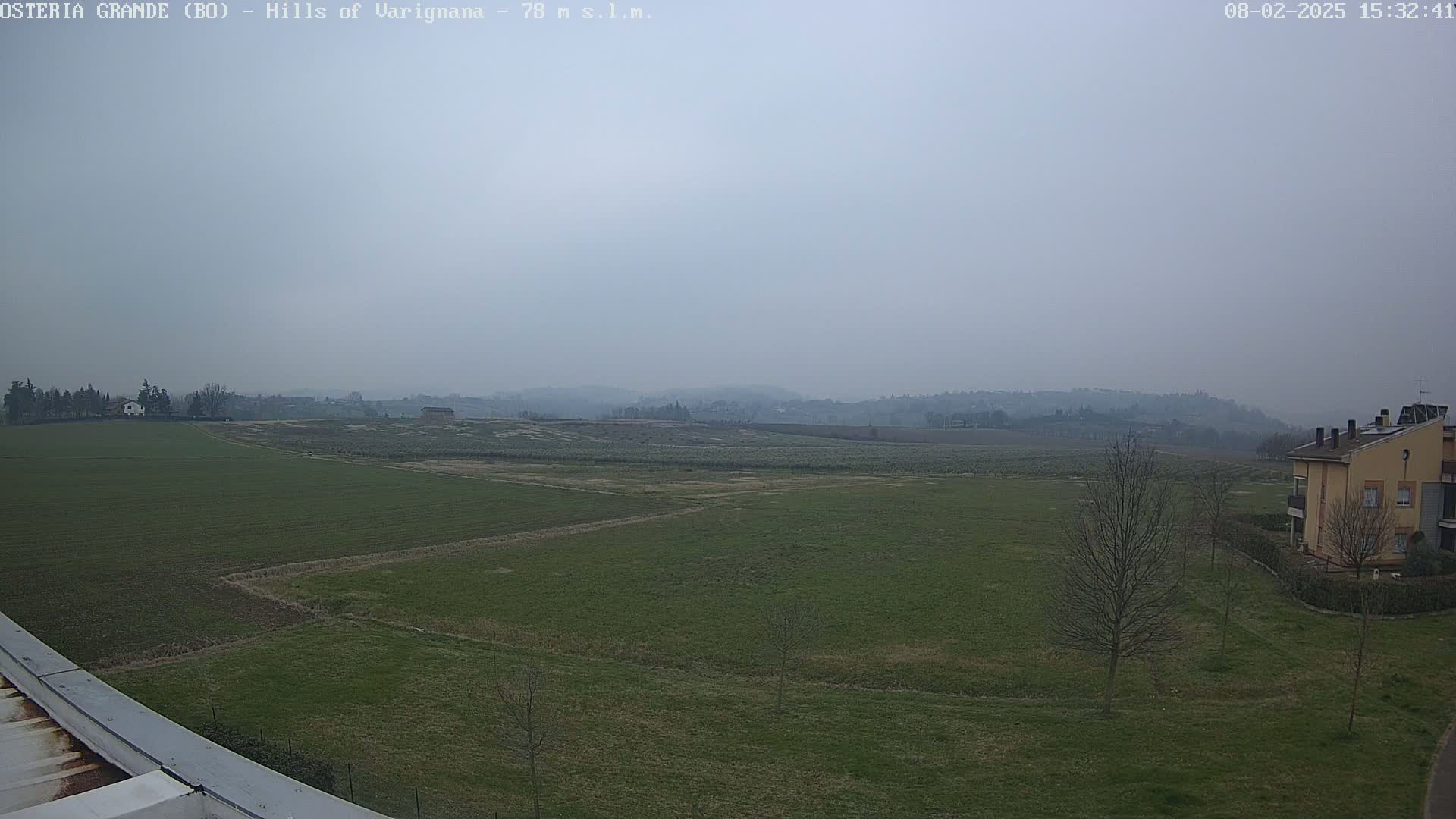 A hazy view of a flat, green field stretches to rolling hills in the distance under a gray sky.