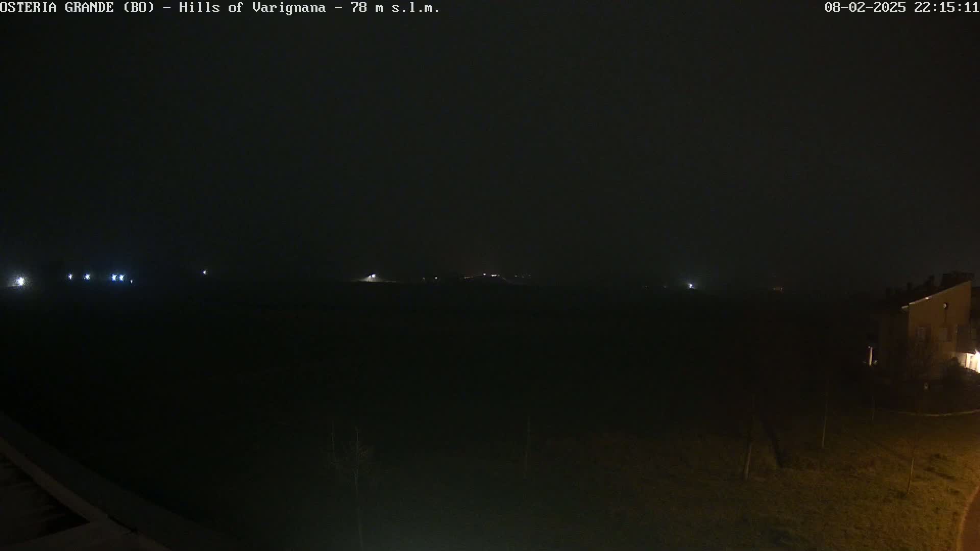 A dark, nighttime scene shows a dimly lit landscape with a few distant lights visible under foggy or hazy conditions.