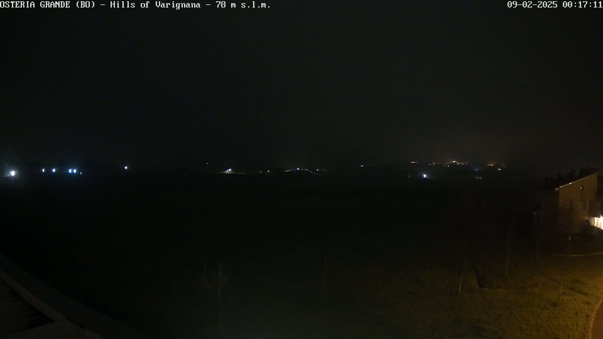 A dark, nighttime view of a flat landscape with a few distant lights and a building partially visible on the right.