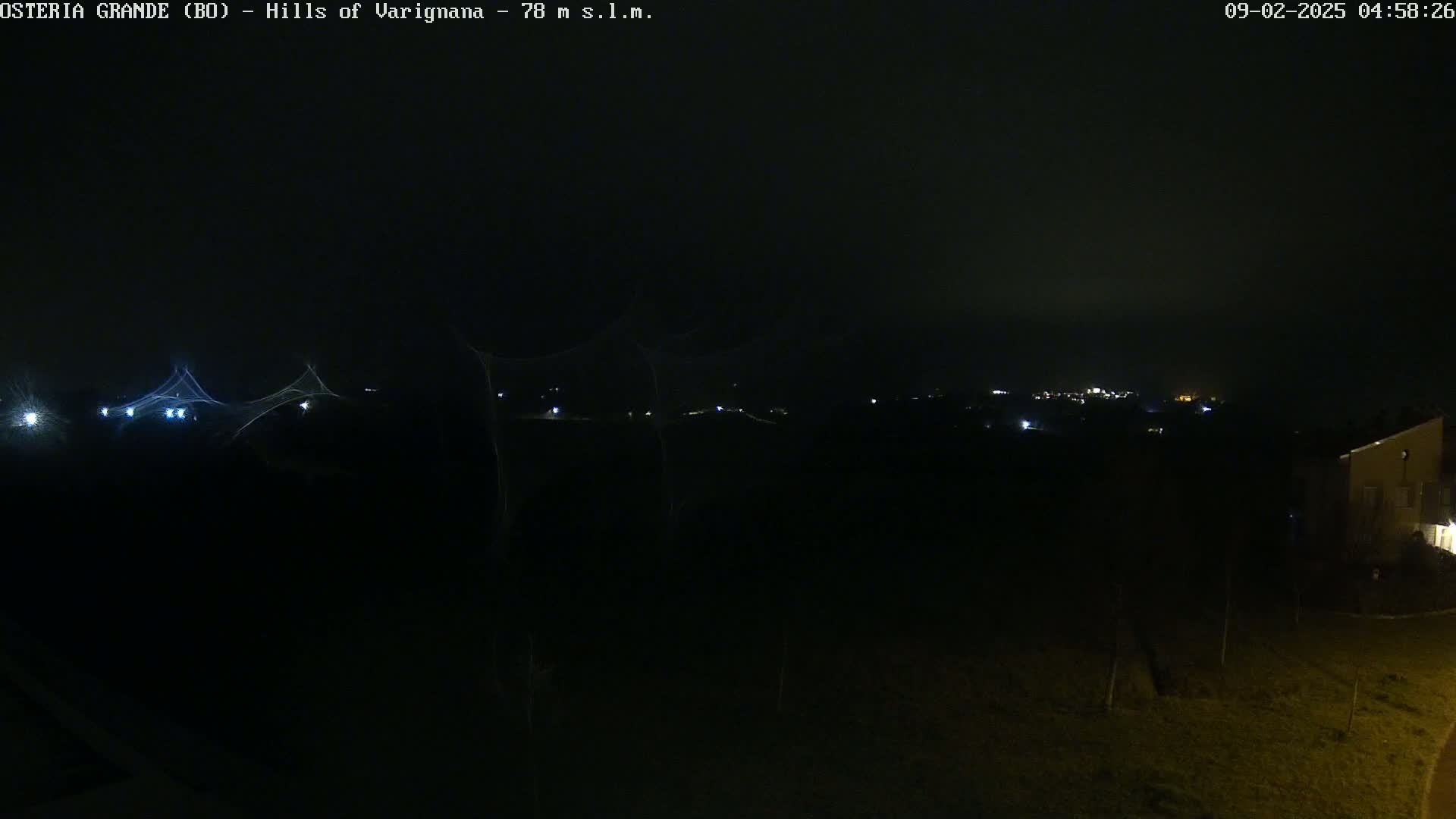 A nighttime, dark, and slightly hazy view of a distant, dimly lit town from a slightly elevated position, with a few lights visible in the foreground and a building on the lower right.