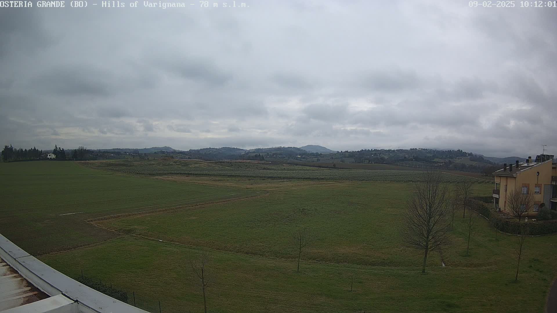 A wide, flat expanse of green fields stretches out under a mostly cloudy sky, with distant hills visible in the background and a few buildings near the bottom right.