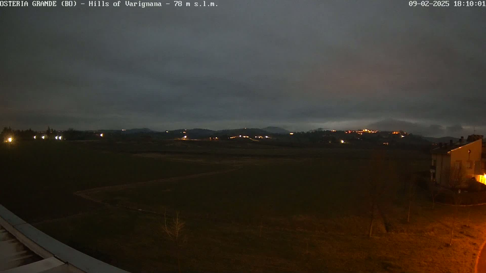 A dark, overcast night scene shows a flat, dark field with a few distant lights on a hill in the background and a building to the right.