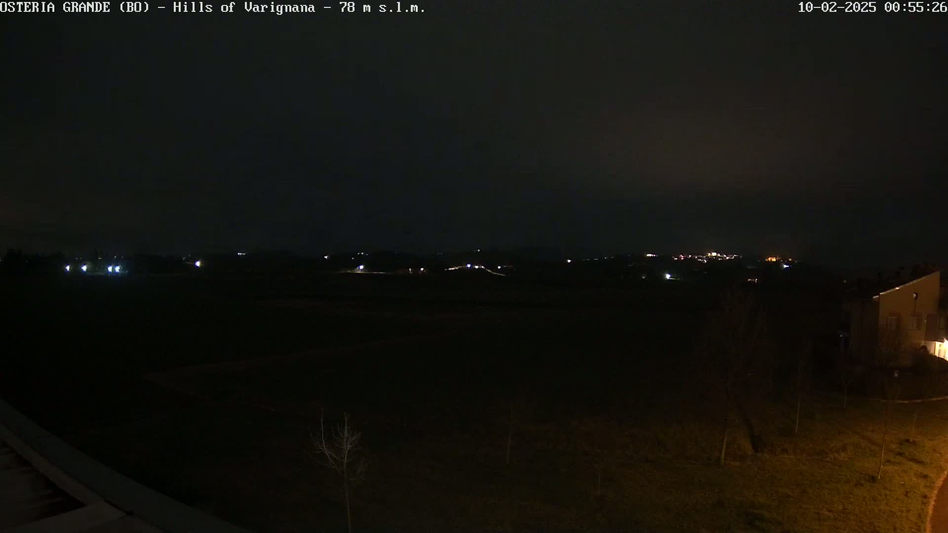 A dark nighttime landscape shows a few distant lights in the hills and a building on the right, under a clear, dark sky.
