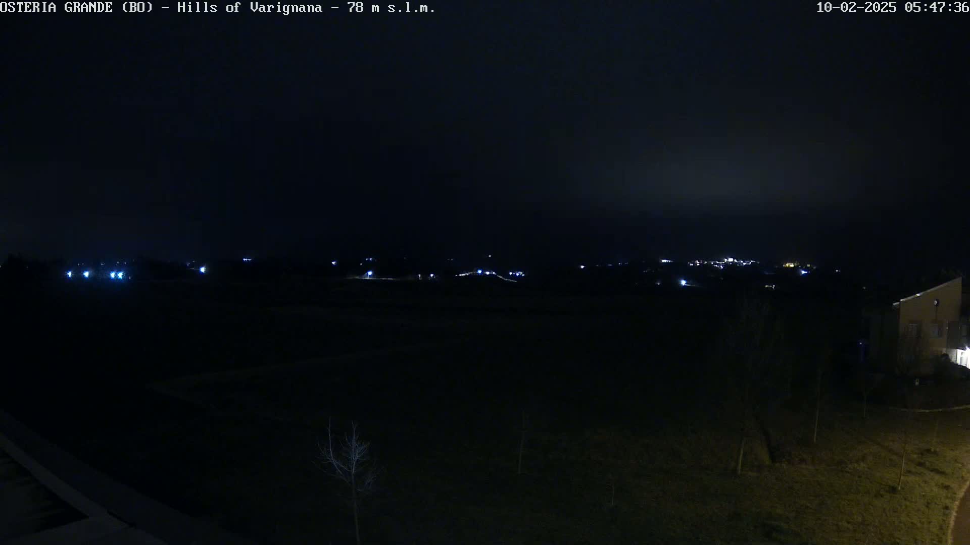 A nighttime, dark view of a mostly flat, sparsely vegetated landscape with a few distant lights and a building visible in the lower right corner.