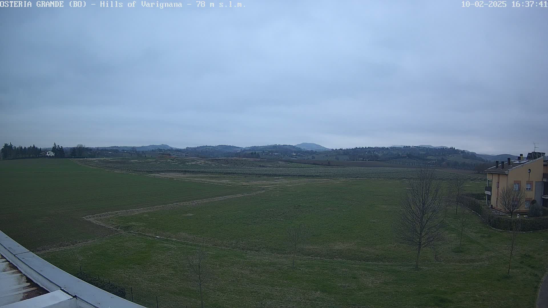 A view of a flat, green field with a few trees and buildings in the foreground, rolling hills in the background, under an overcast sky.