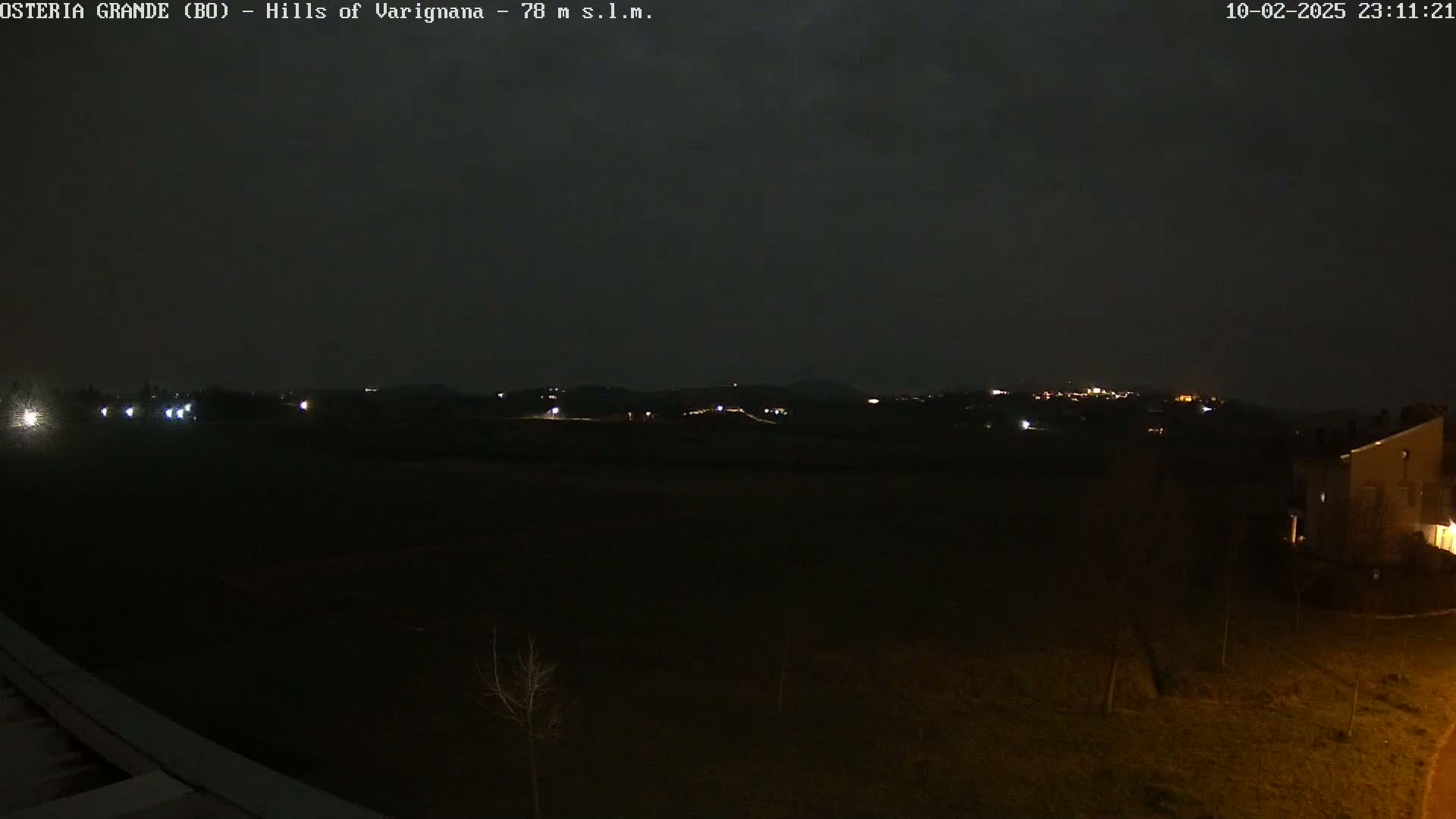 A nighttime view of dimly lit hills in the distance, with a few lights visible in the foreground and a dark, overcast sky.