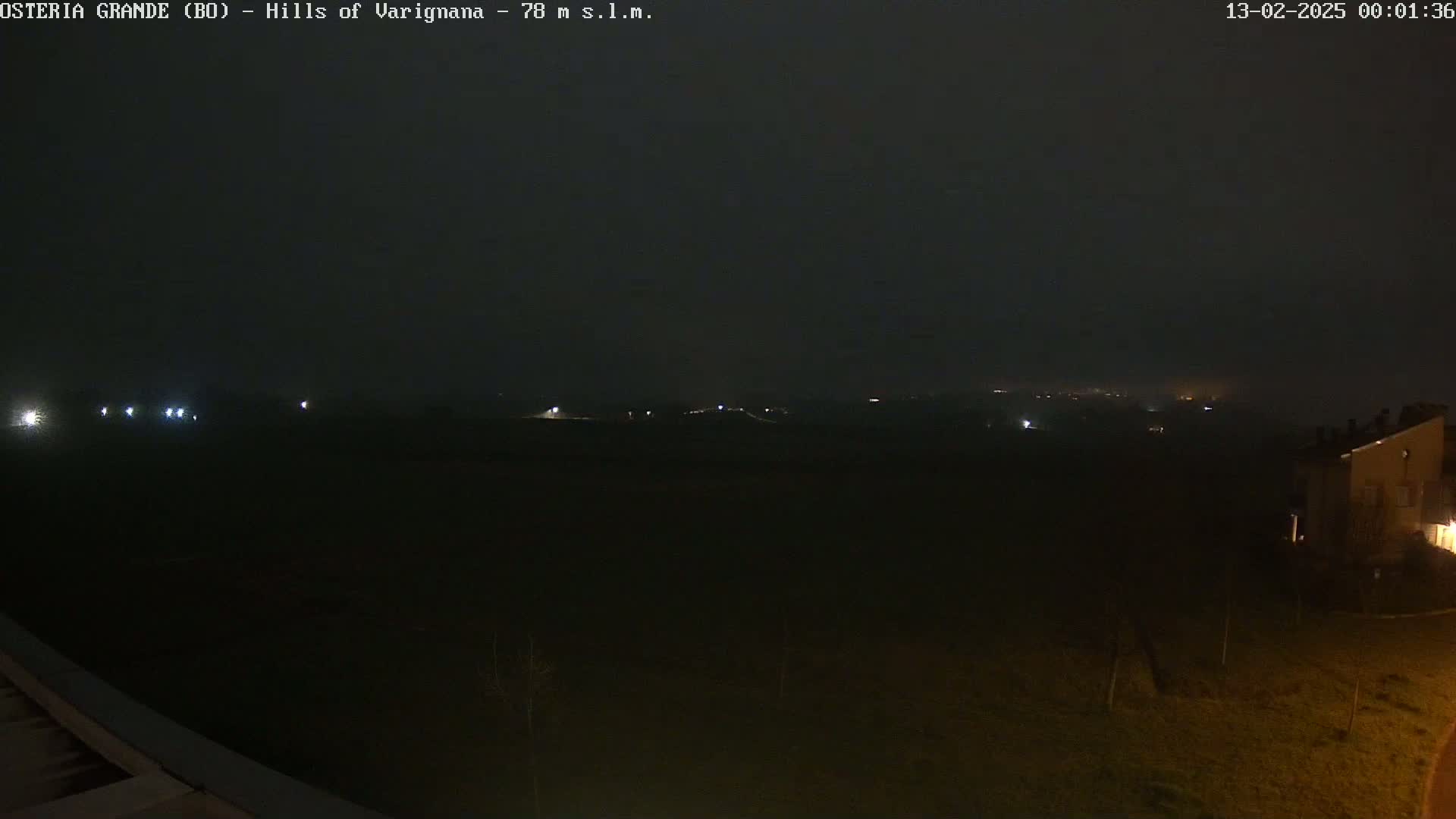 A nighttime, foggy view shows a dark landscape with a few distant lights and a building partially visible in the lower right corner.
