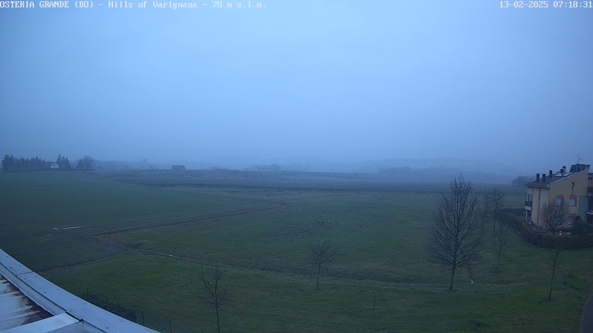 A hazy, pale blue sky hangs over a flat expanse of green fields and a few bare trees, with a building visible in the lower right corner.