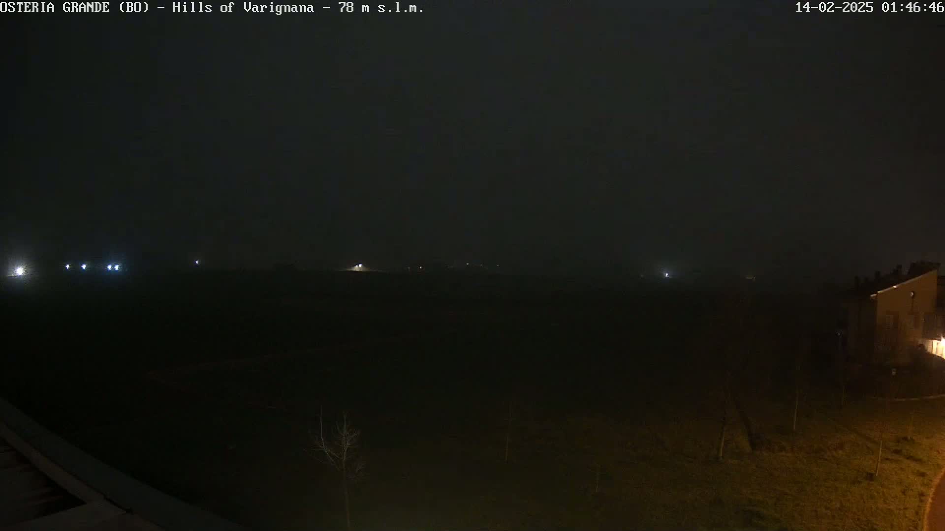 A dark, foggy nighttime scene shows a flat expanse of land with a few distant lights, and a building partially visible in the bottom right corner.