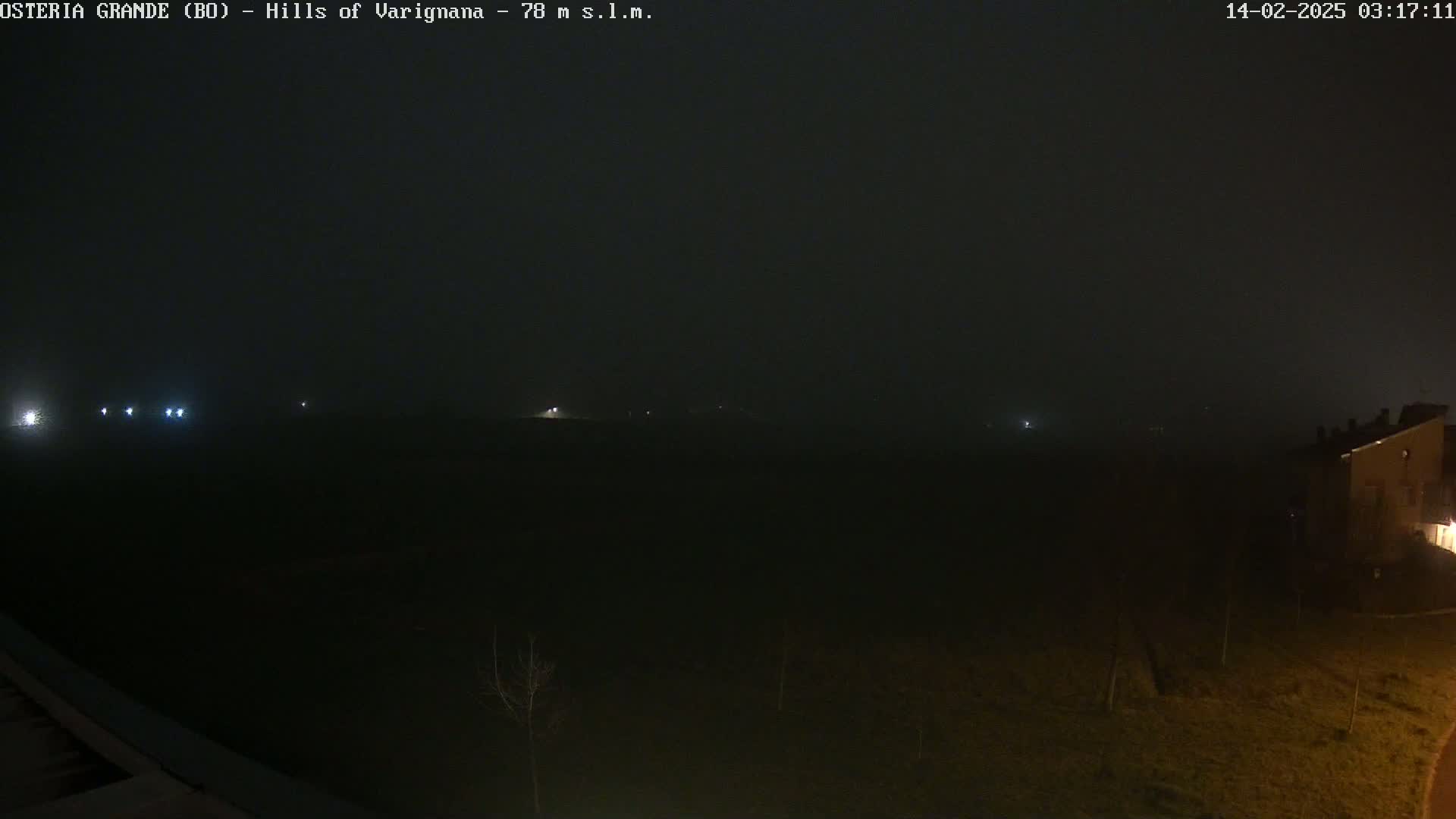 A dark, foggy night scene shows dimly lit hills and a few distant lights, with a building partially visible in the lower right corner.