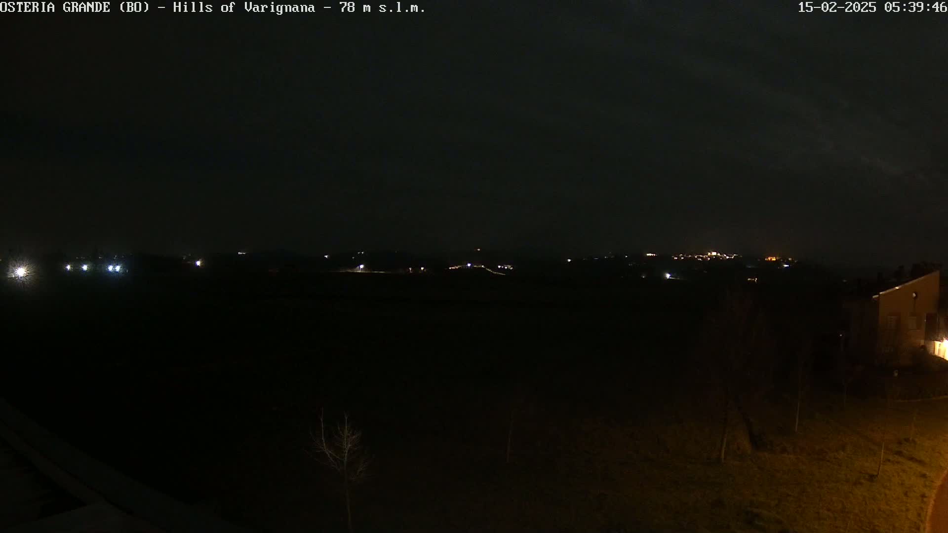 A dark, nighttime view of distant, dimly lit hills and a few nearby buildings under a cloudy sky.