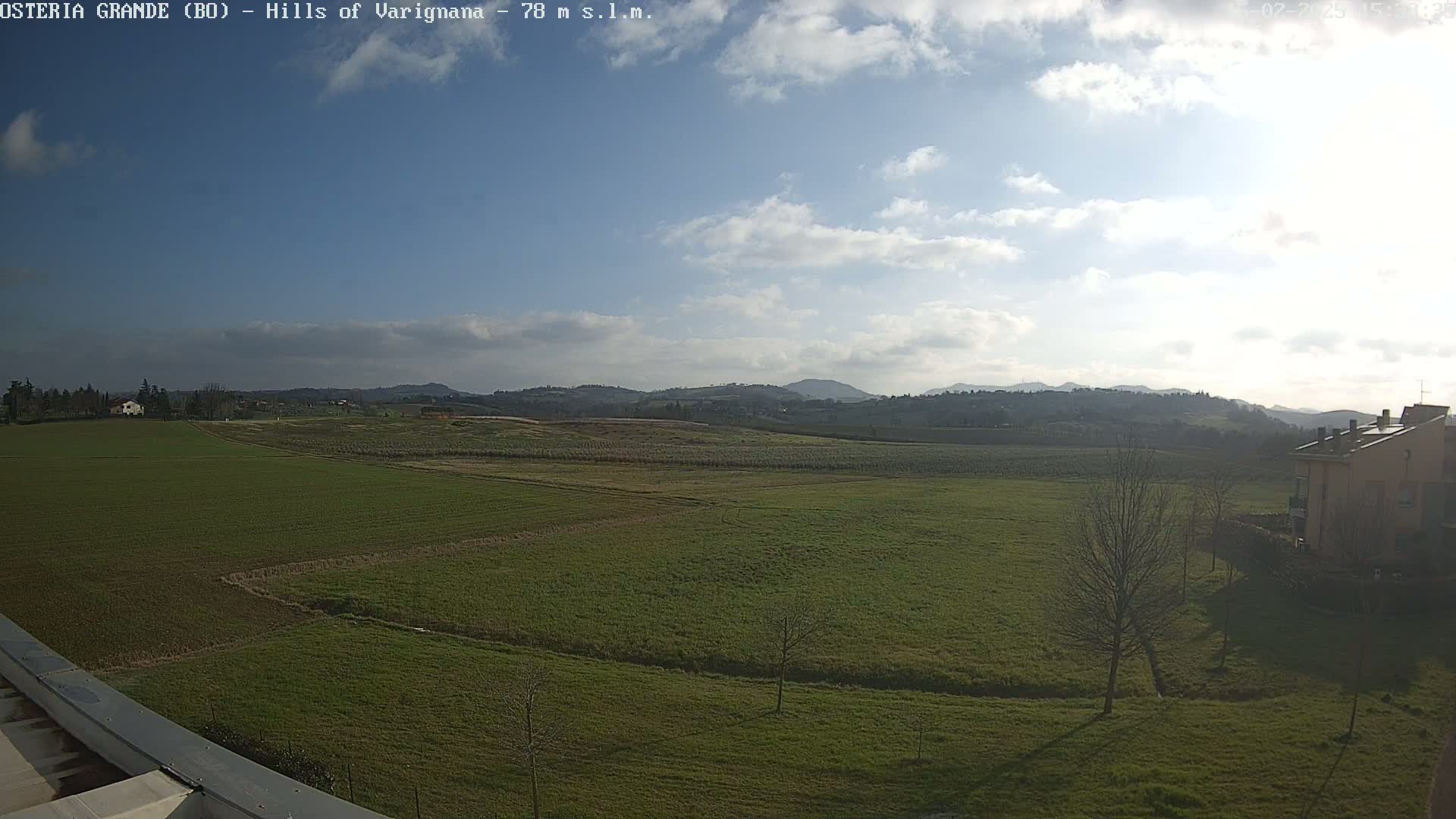 A partly cloudy day reveals a wide expanse of green farmland, dotted with a few buildings and trees, stretching towards distant hills.
