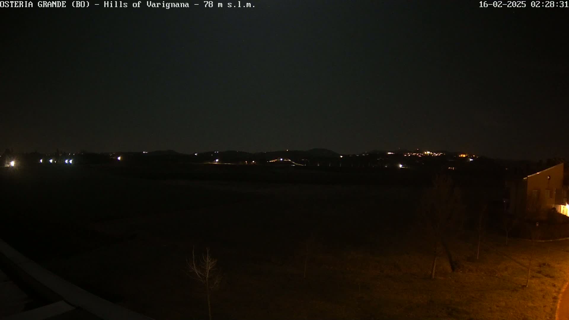 A nighttime, dark view of dimly lit hills in the distance, with a flat, dark foreground containing sparsely placed trees and a building.