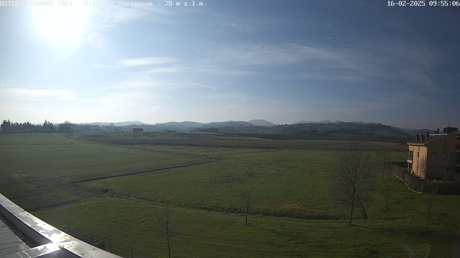 A sunny day reveals a sprawling green field, dotted with a few bare trees, extending to distant rolling hills under a mostly clear blue sky.