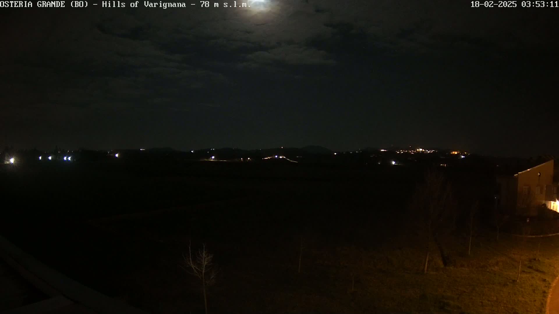 A nighttime view of dimly lit hills and a few buildings under a partly cloudy moonlit sky.