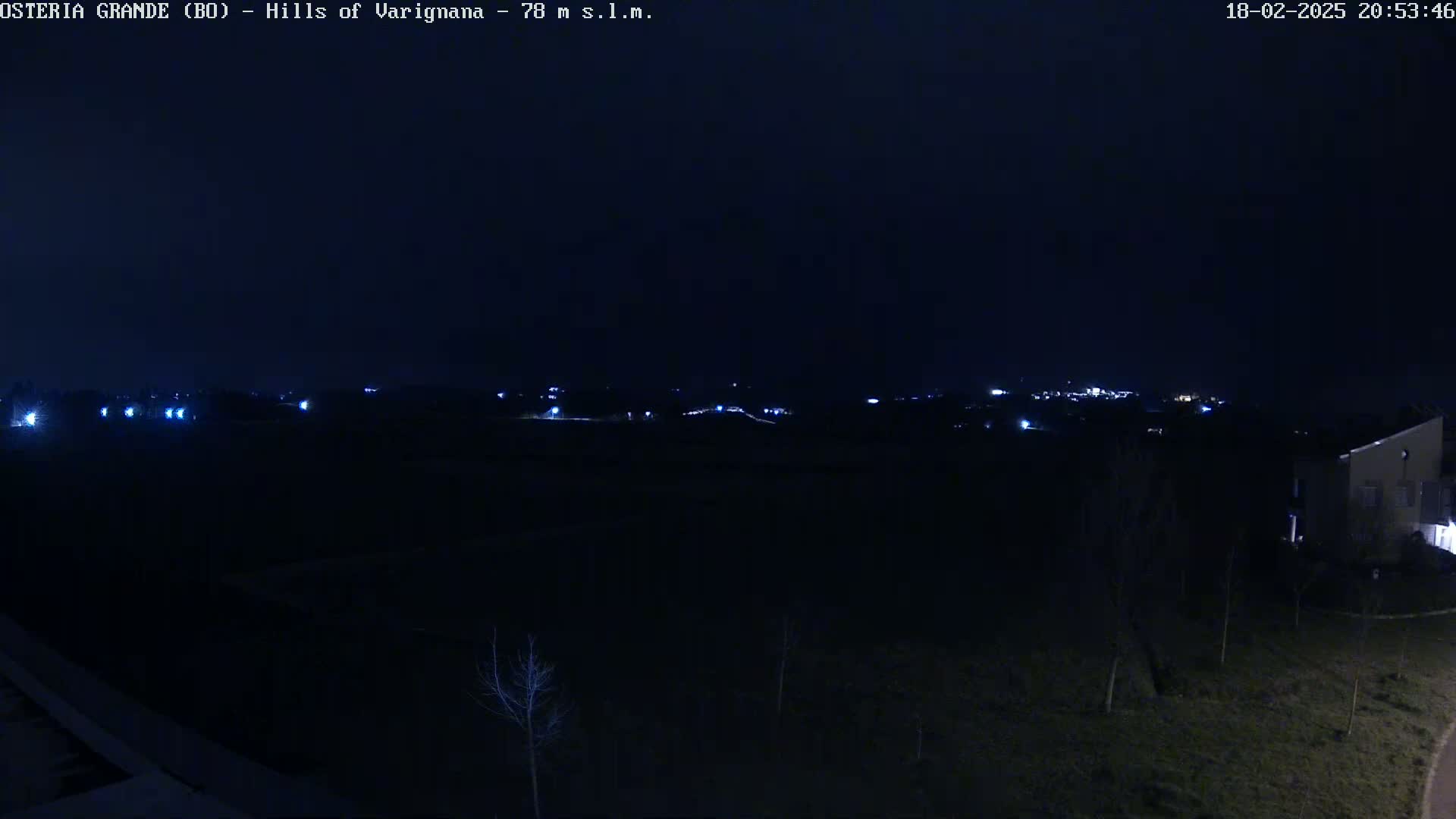 A nighttime, long shot of a dark landscape with a few distant lights and a building visible in the lower right corner.