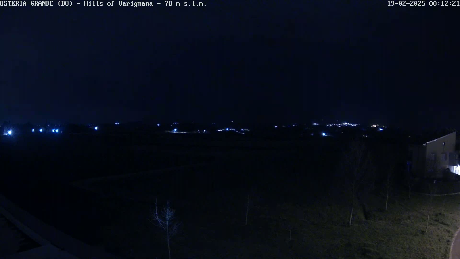 A dark, nighttime view of sparsely lit hills and a few buildings in the distance under a clear sky.