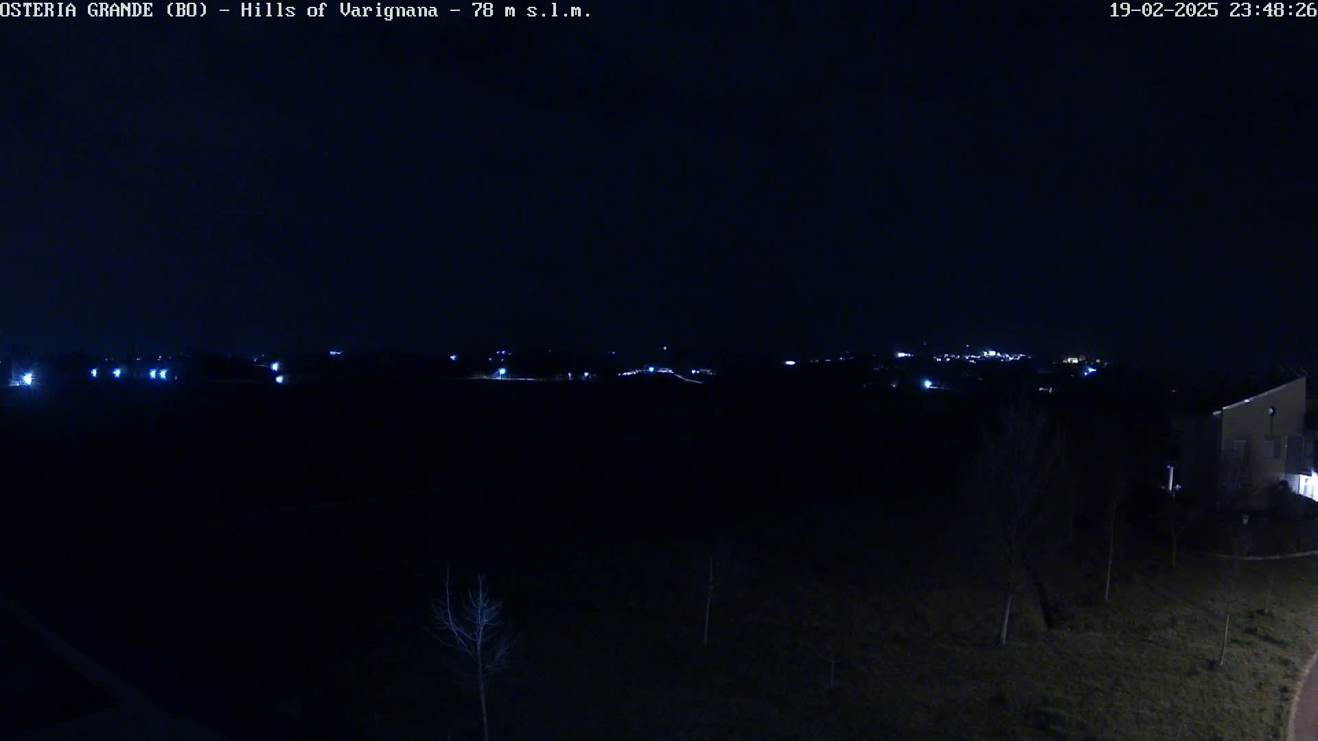 A nighttime view of a dark landscape with a few distant lights and a building in the bottom right corner.