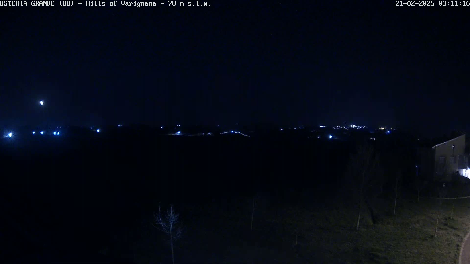 A nighttime, dark landscape shows a distant line of lights from a town or village, with a few lights closer to the viewer and a building visible on the lower right.