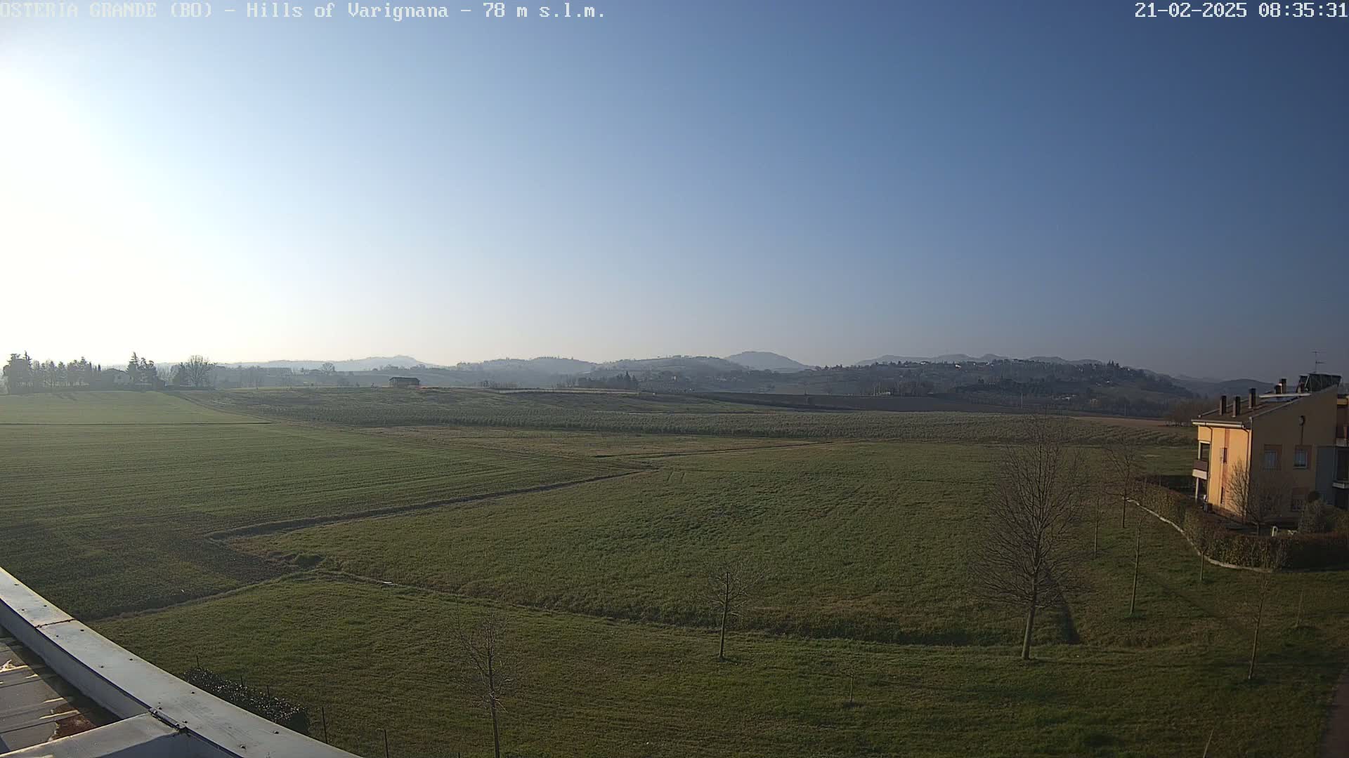 A vast, flat field of green grass stretches towards distant, hazy hills under a clear, bright blue sky.