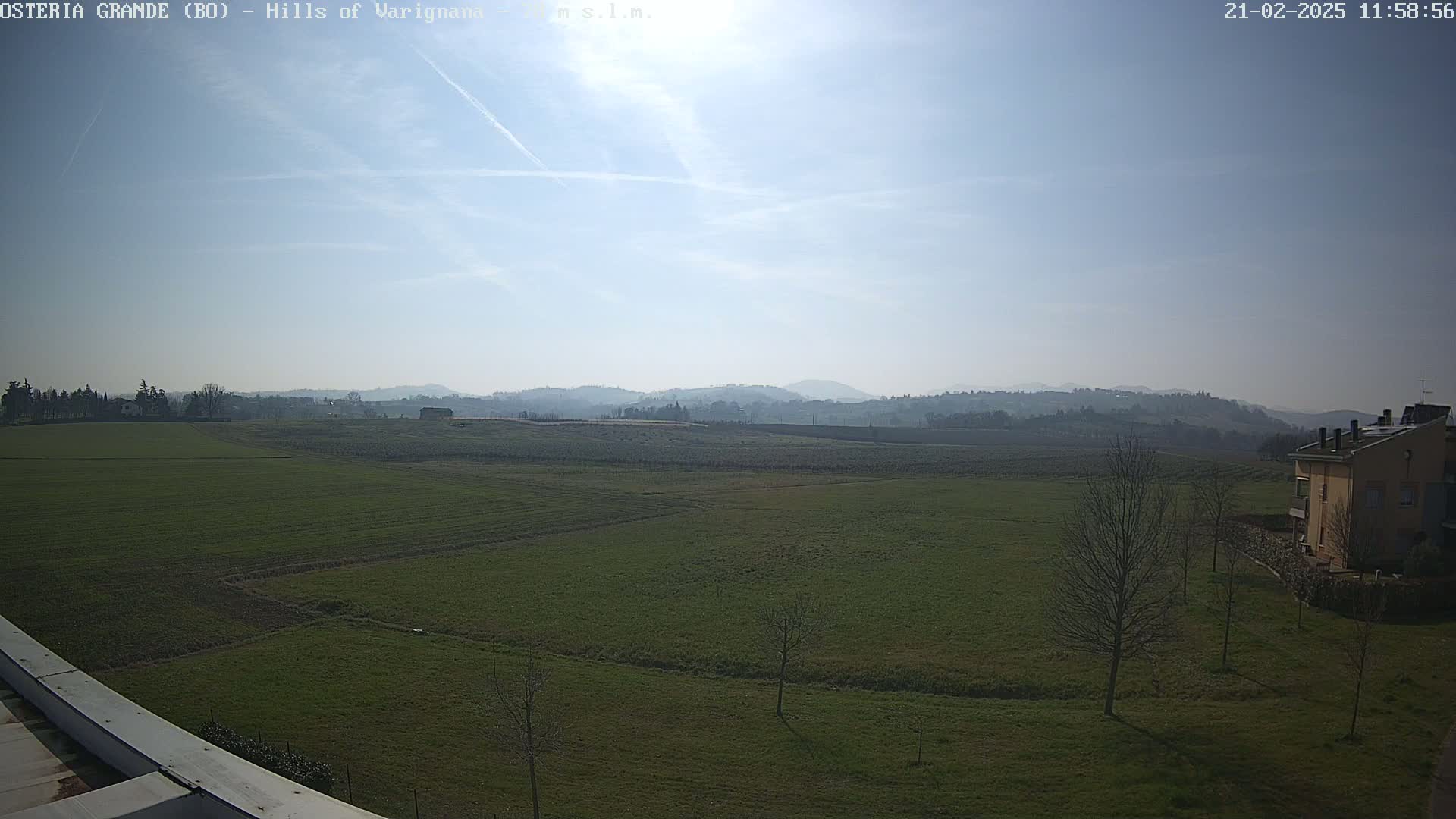 A sunny day reveals a wide expanse of green fields, dotted with a few bare trees, leading to rolling hills in the distance.