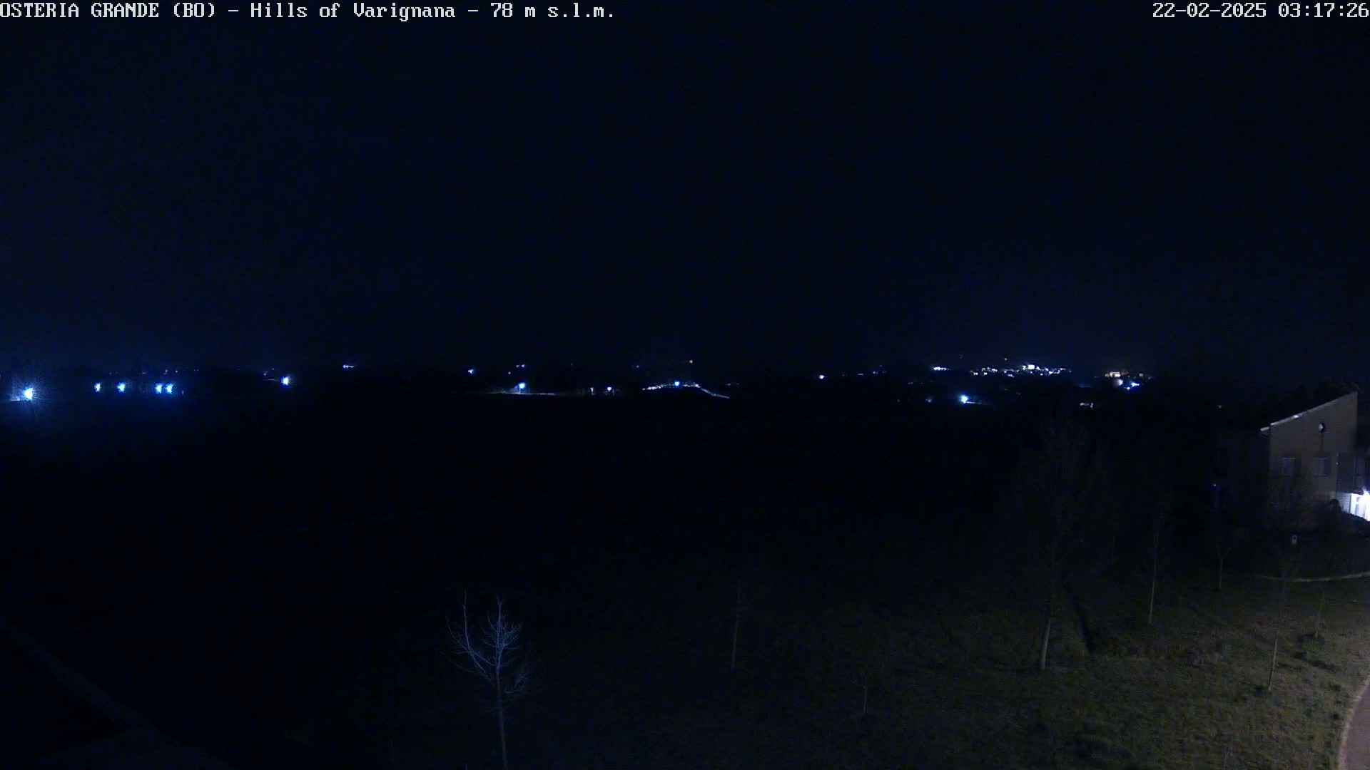 A nighttime, dark landscape shows a distant town illuminated by lights, with a few lights closer in the foreground, and a building partially visible to the right.